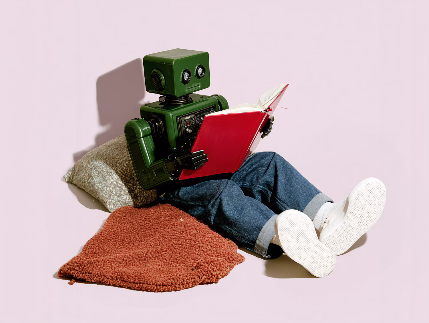 Robot verde con jeans y tenis blancos leyendo un libro rojo, sentado entre cojines sobre un fondo rosa claro.